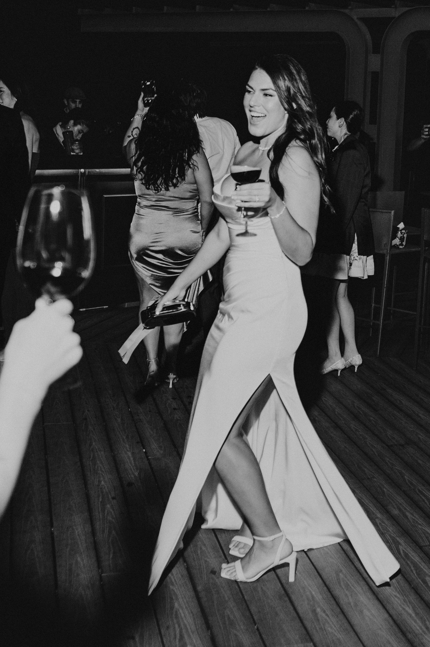 Black and white photo of woman in slit gown dancing with wine glass at wedding reception.