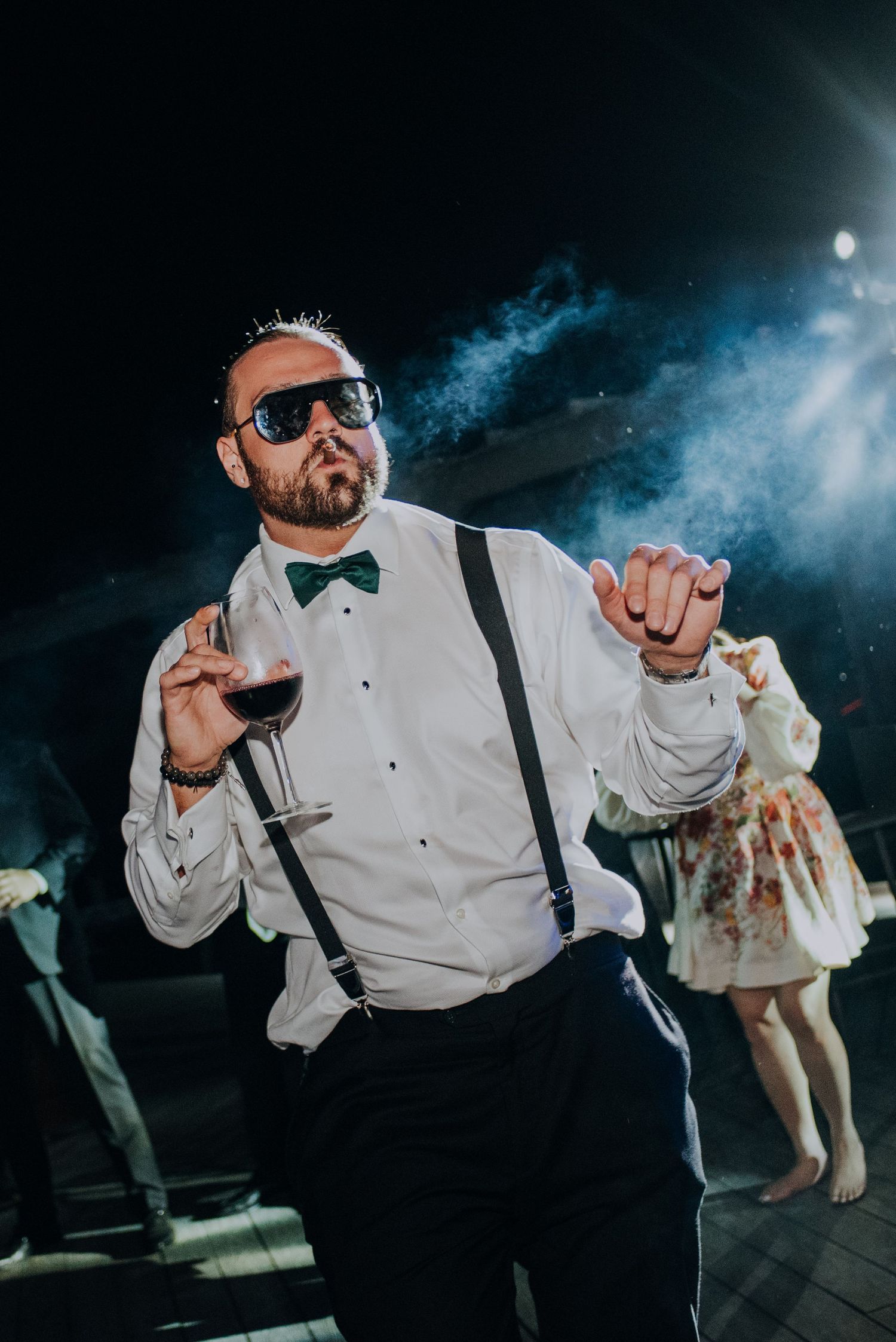 Stylish wedding guest in suspenders and bow tie dancing with sunglasses and red wine at reception.