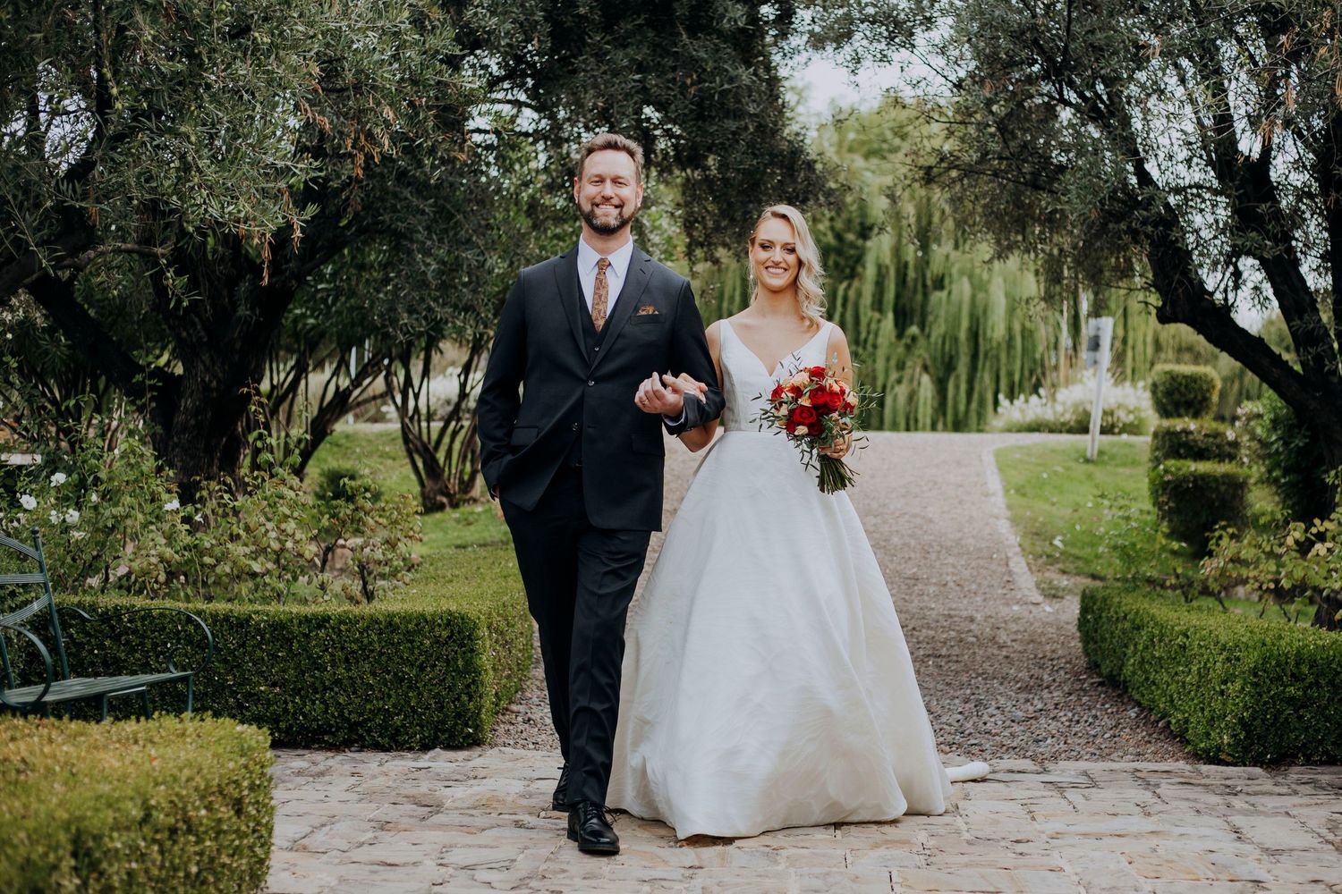 Bride and groom walking hand in hand through lush garden pathway.