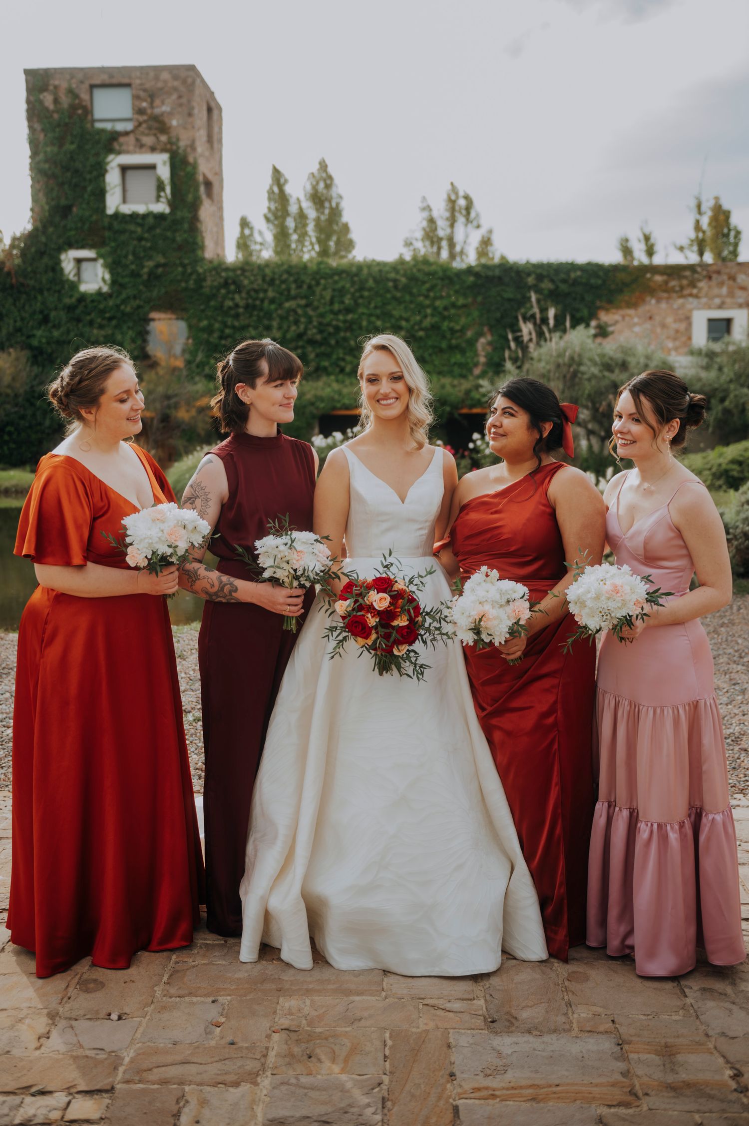 Bride in white gown poses with four bridesmaids in red and pink dresses holding bouquets outdoors.