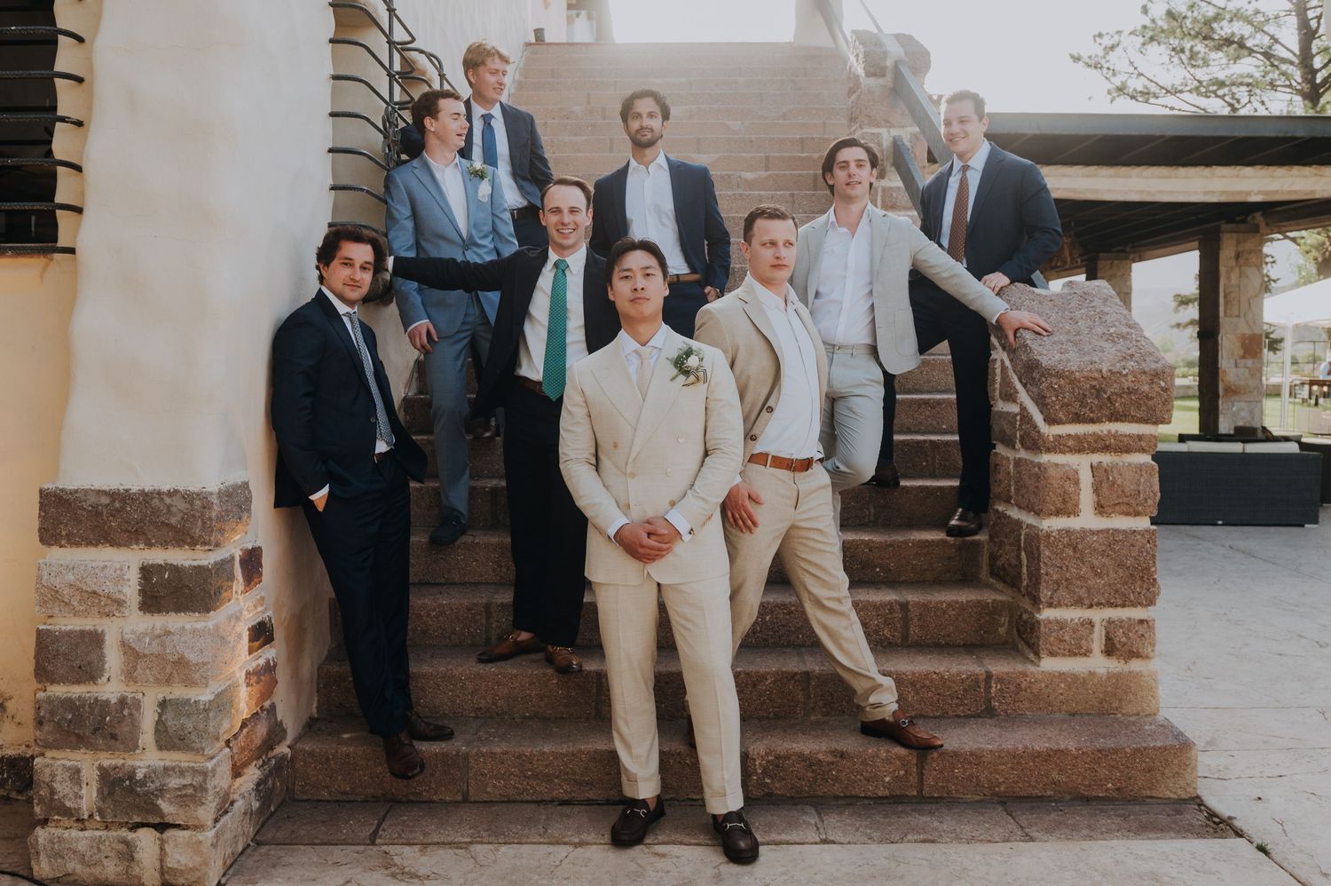 Groom in cream suit poses with groomsmen in suits on stone steps at outdoor wedding venue.