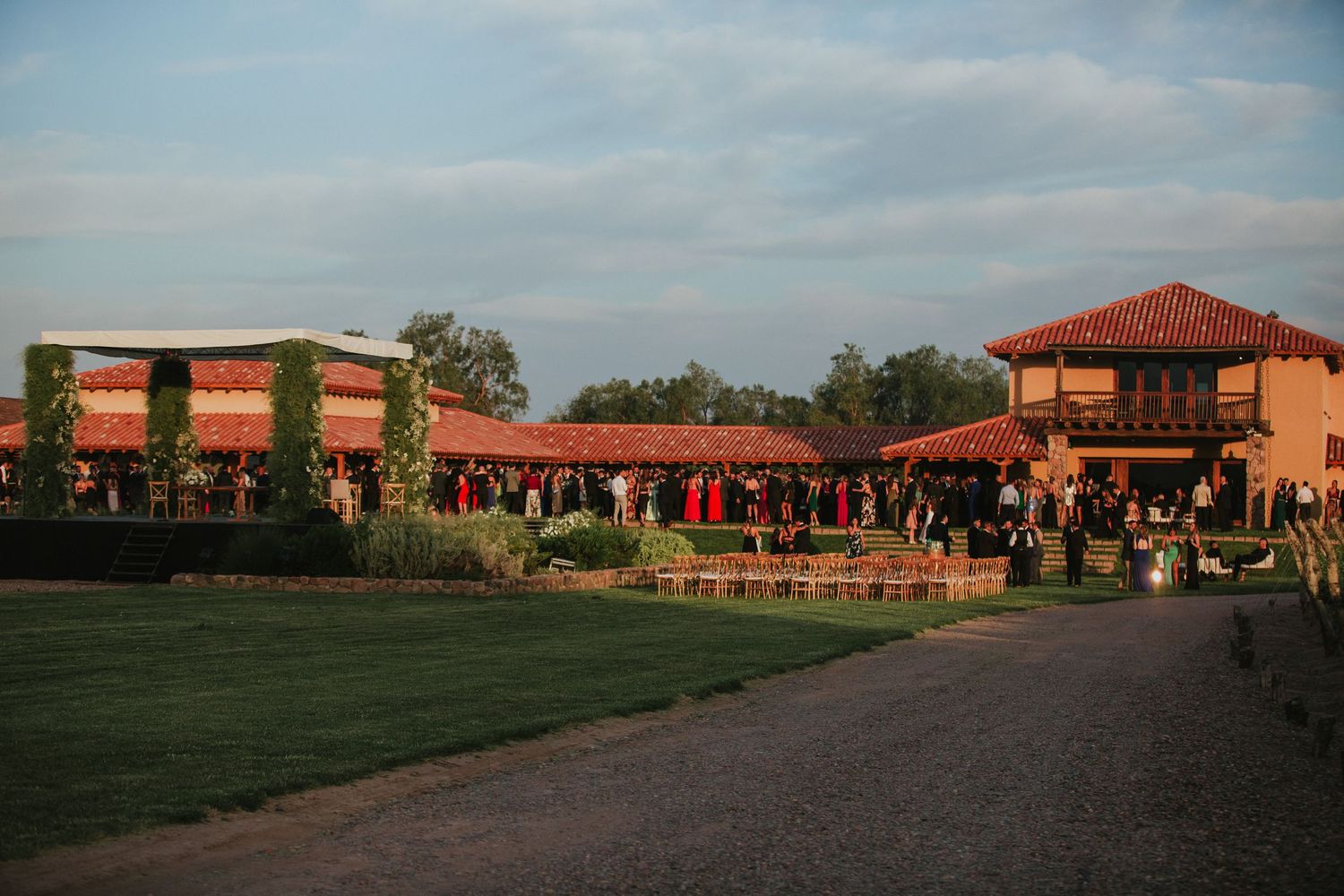 Outdoor wedding reception at a rustic venue with guests gathered on a lawn at golden hour.