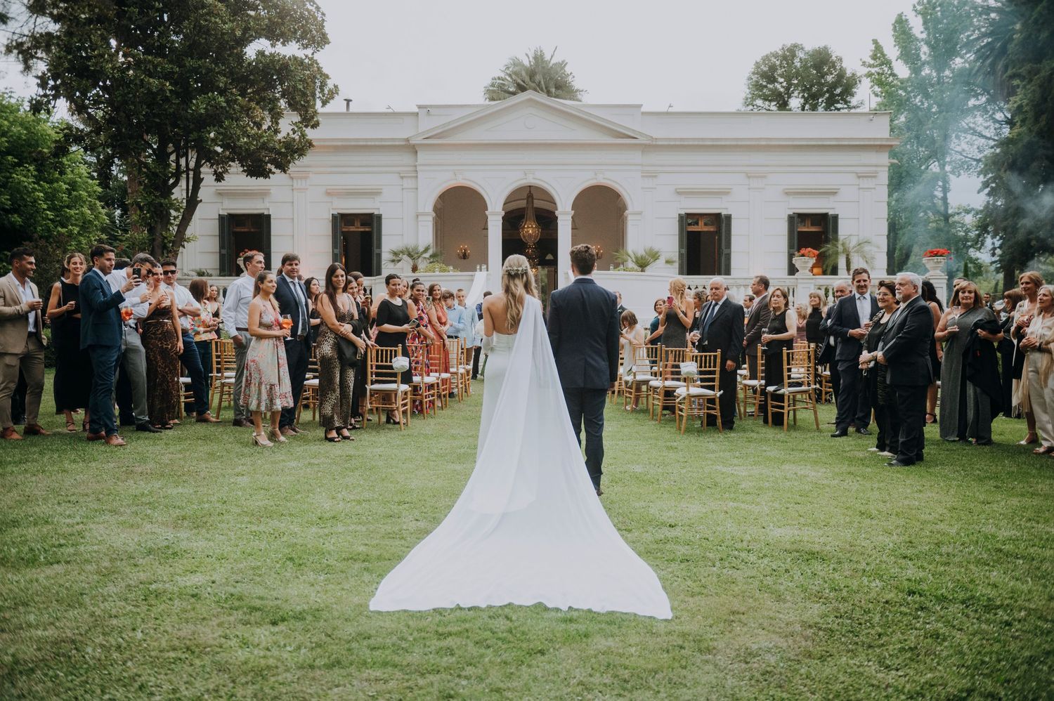 Bride and groom walk down outdoor aisle at elegant white venue surrounded by guests on a lush green lawn.
