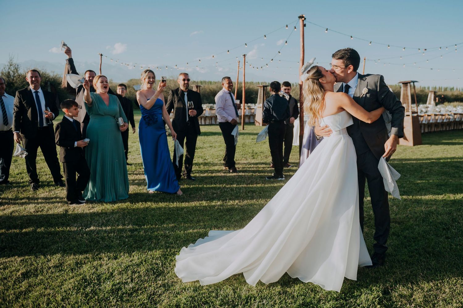 Bride and groom share romantic kiss while wedding guests celebrate on green lawn.