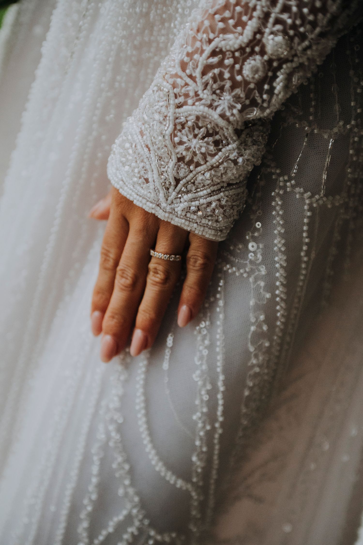 Close-up of a bride's hand wearing diamond rings on an intricate beaded lace wedding gown.