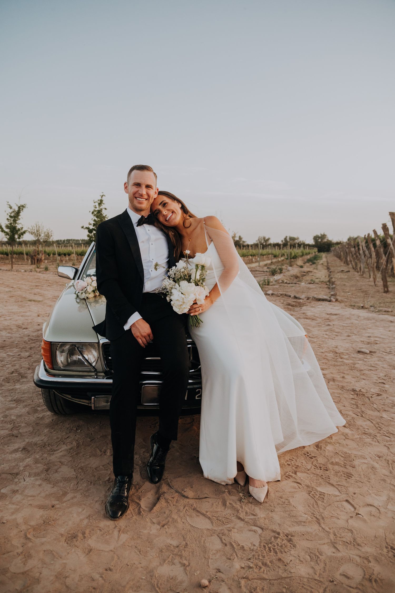 Bride and groom leaning against vintage car in sandy vineyard at golden hour.