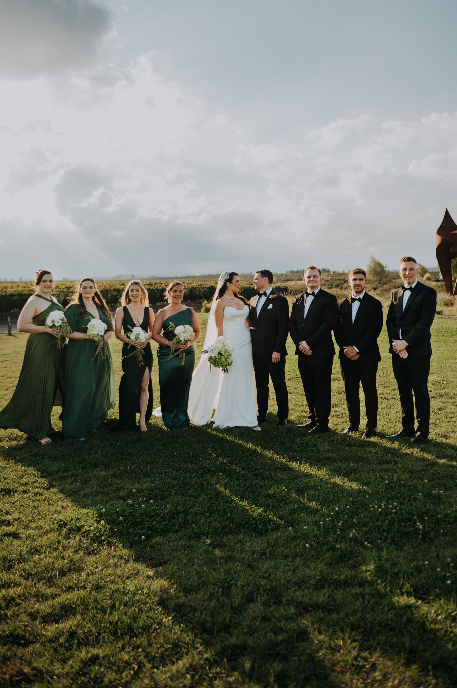 Wedding party poses outdoors on green grass, bride in white gown with bridesmaids in emerald green dresses and groomsmen in black tuxedos.