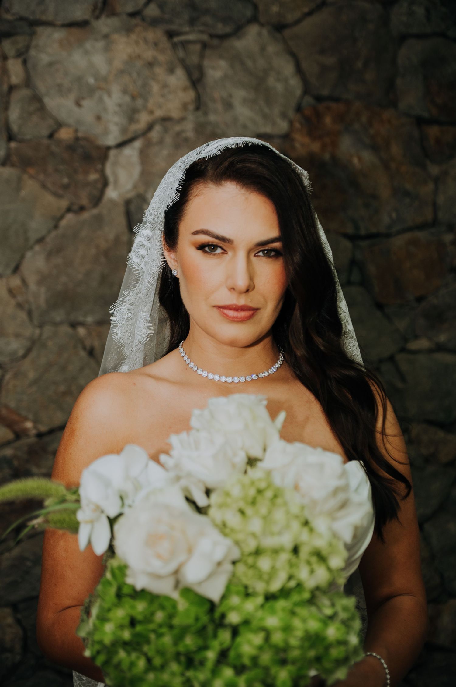 Beautiful bride in lace veil holds white and green hydrangea bouquet against stone wall.
