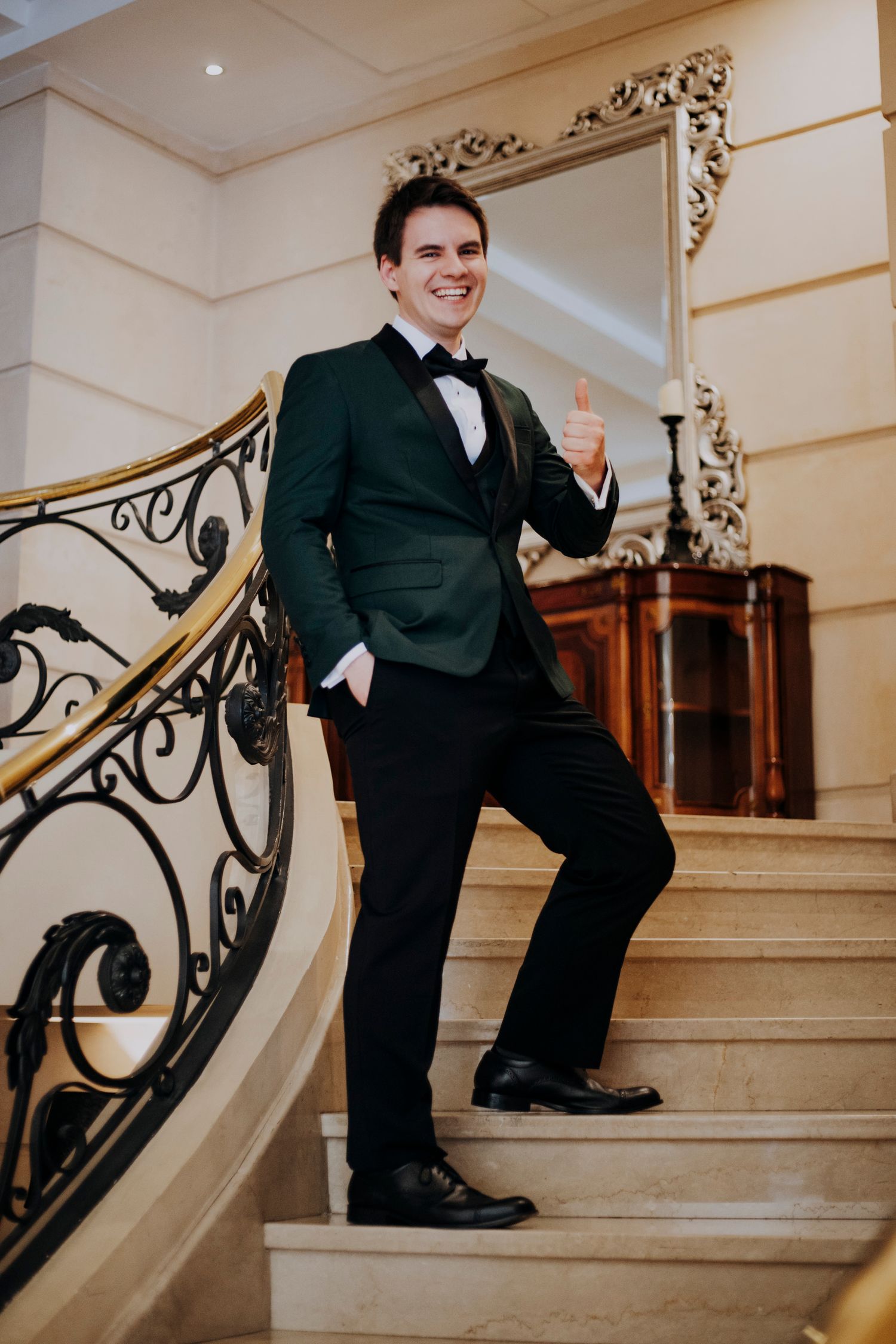 Smiling groom in green tuxedo jacket giving thumbs up on elegant staircase.