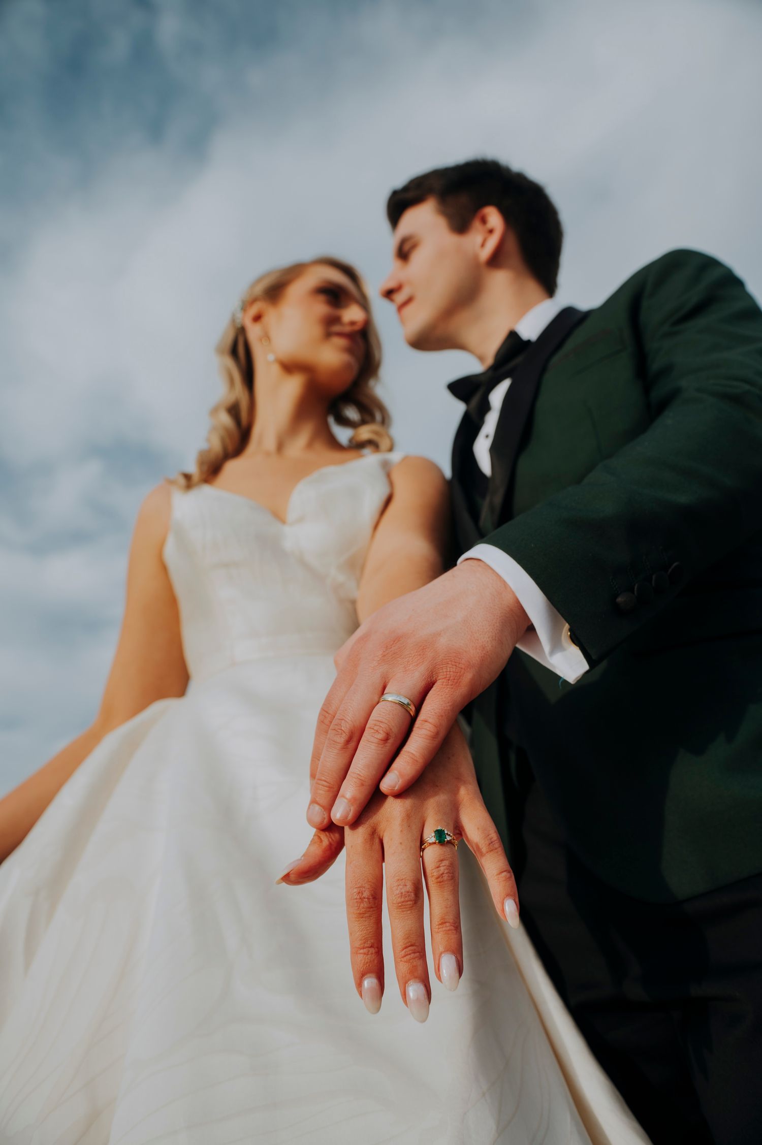 Newlyweds display wedding rings to camera against cloudy sky, bride in white gown groom in green suit.