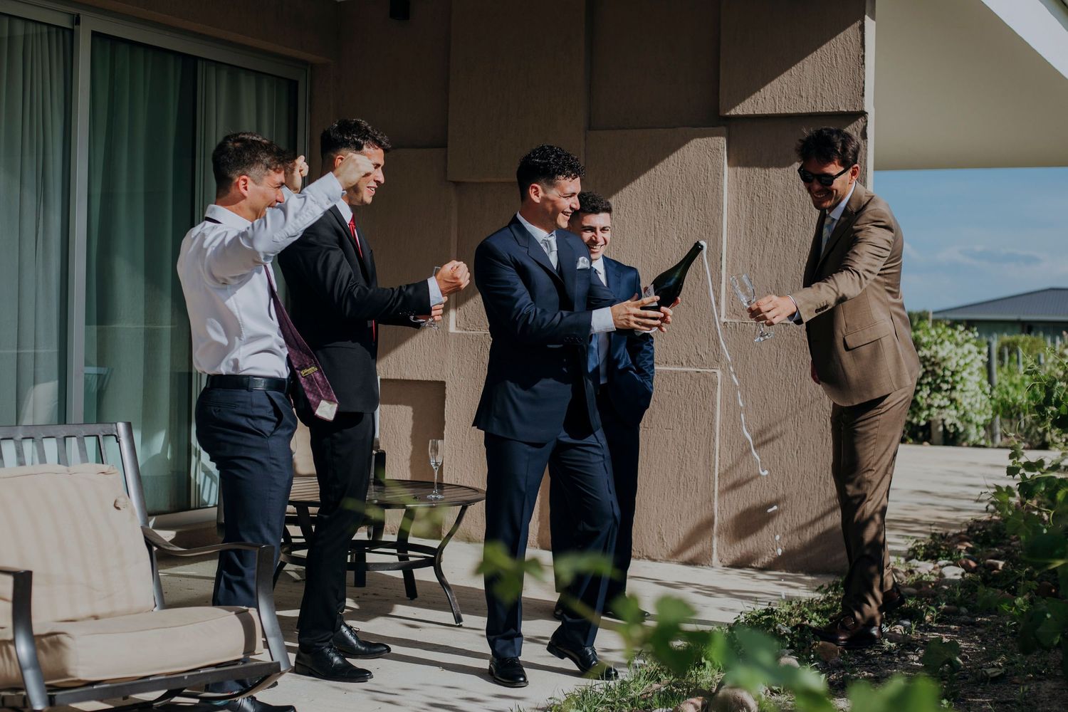 Groomsmen in suits gather on outdoor terrace sharing laughs before wedding ceremony begins.