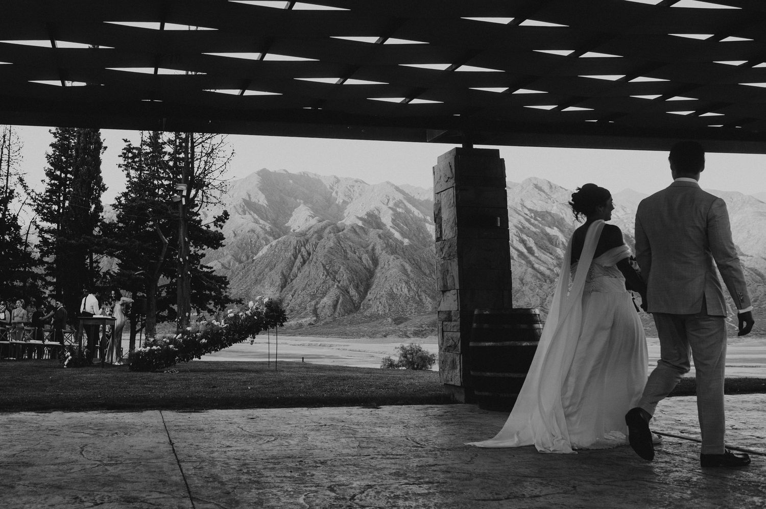 Black and white photo of bride and groom walking under pergola with mountain backdrop.