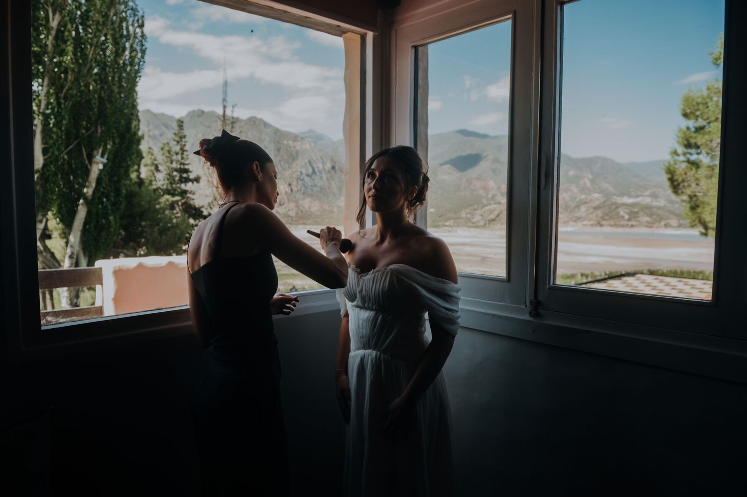 Bride in white off-shoulder gown being helped by bridesmaid near large windows with mountain views.
