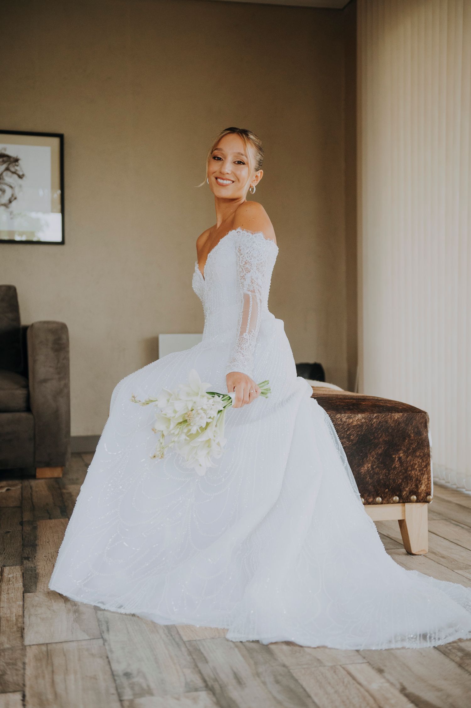 Smiling bride in off-shoulder lace wedding gown holding white floral bouquet while seated indoors.