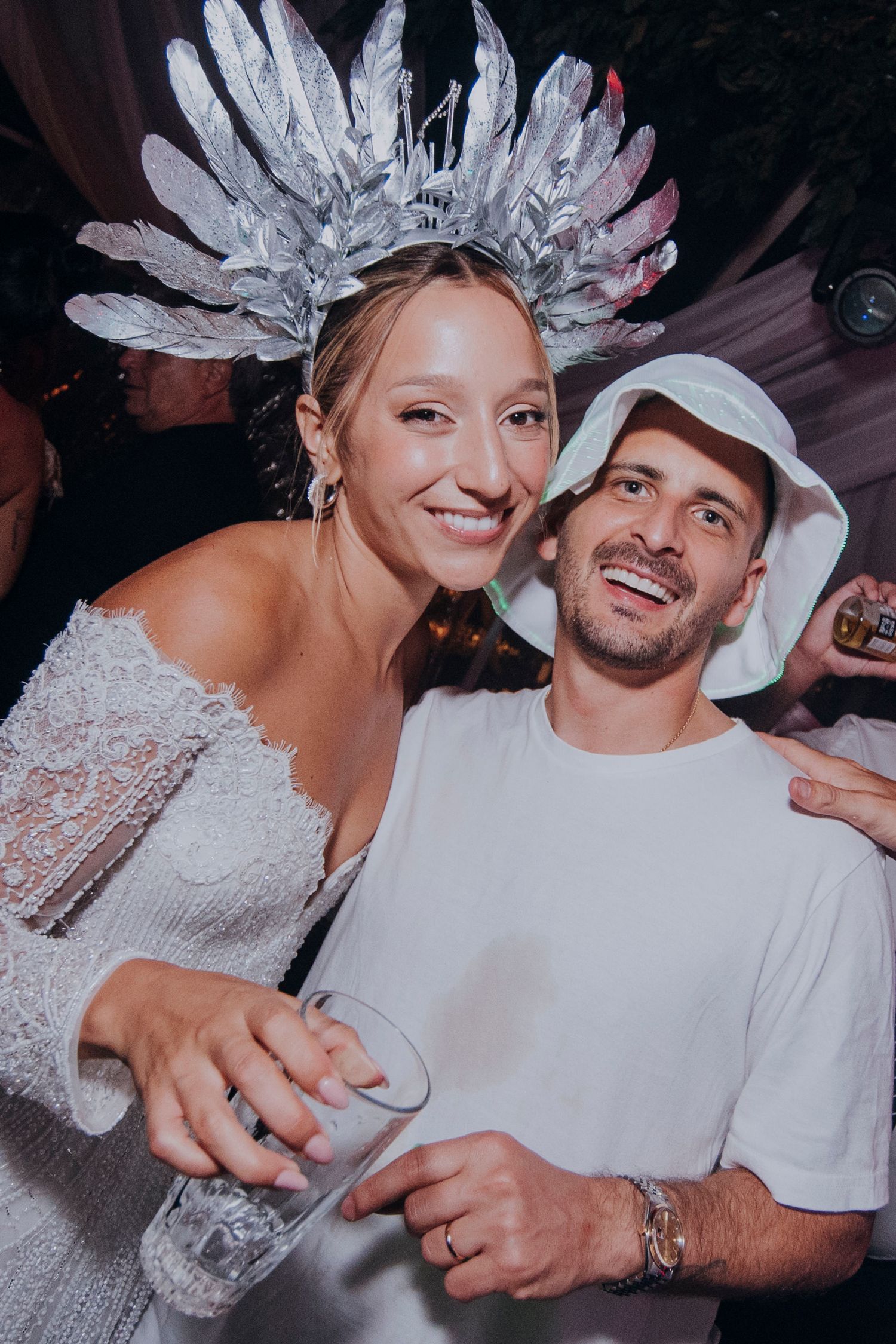 Woman in white dress with silver feather crown and man in white bucket hat smiling at a party.