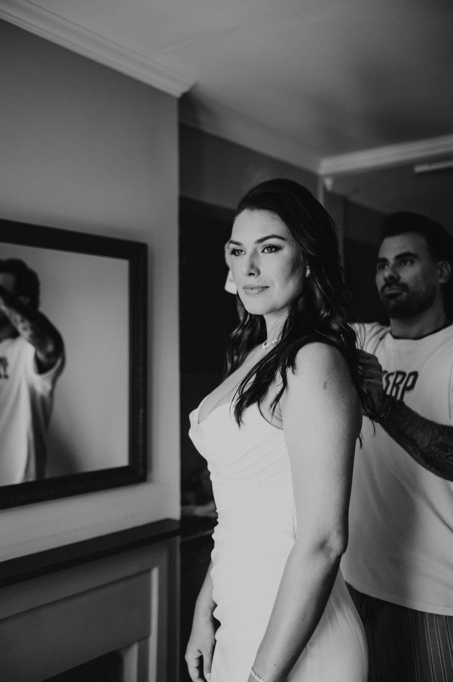 Black and white photo of bride in white dress being helped by groom near a mirror before wedding ceremony.