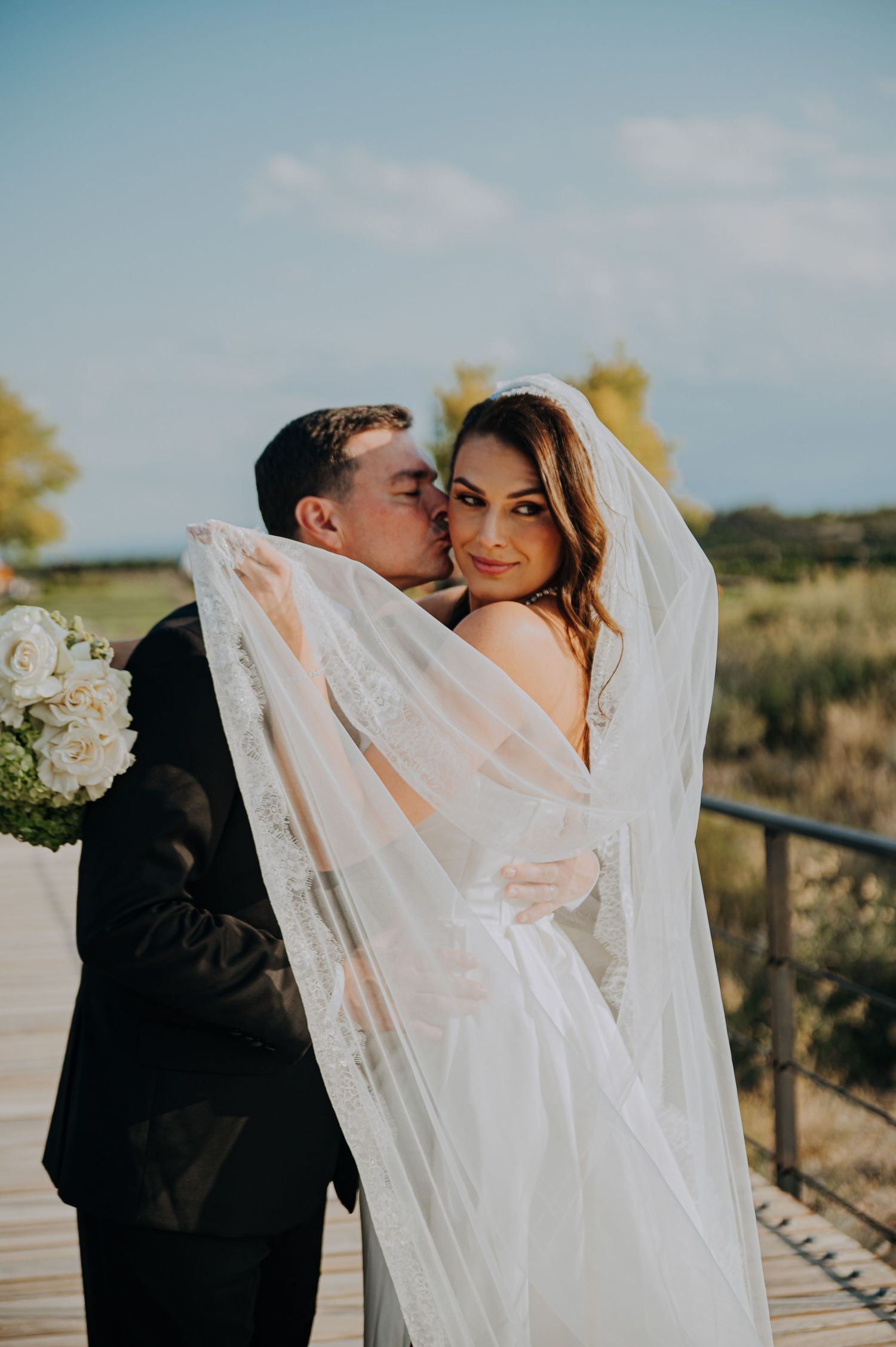 Bride in white gown and veil smiles as groom kisses her cheek on outdoor wooden deck.