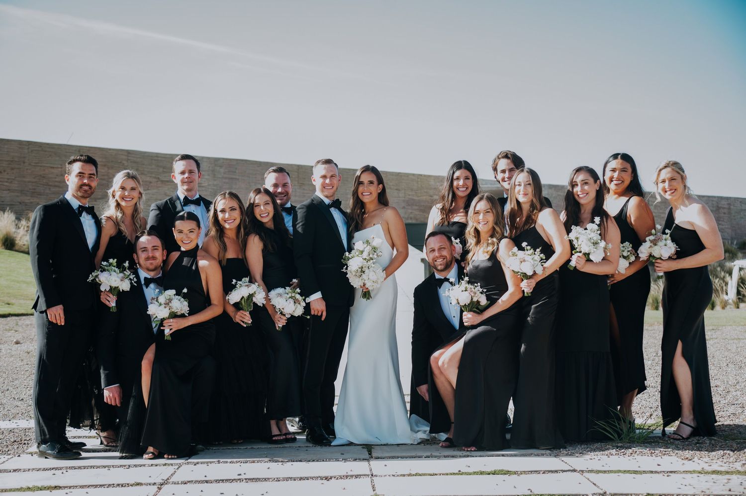 Large wedding party in black formal attire with bride in white gown pose together outdoors at a winery.