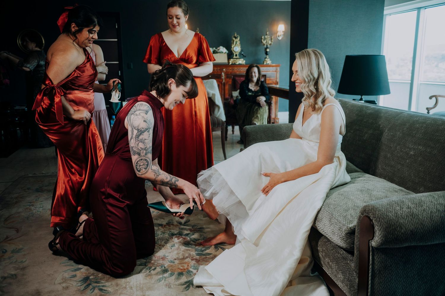 Bridesmaids in red dresses help bride put on shoes while sitting on a couch in a dark-walled bridal suite.