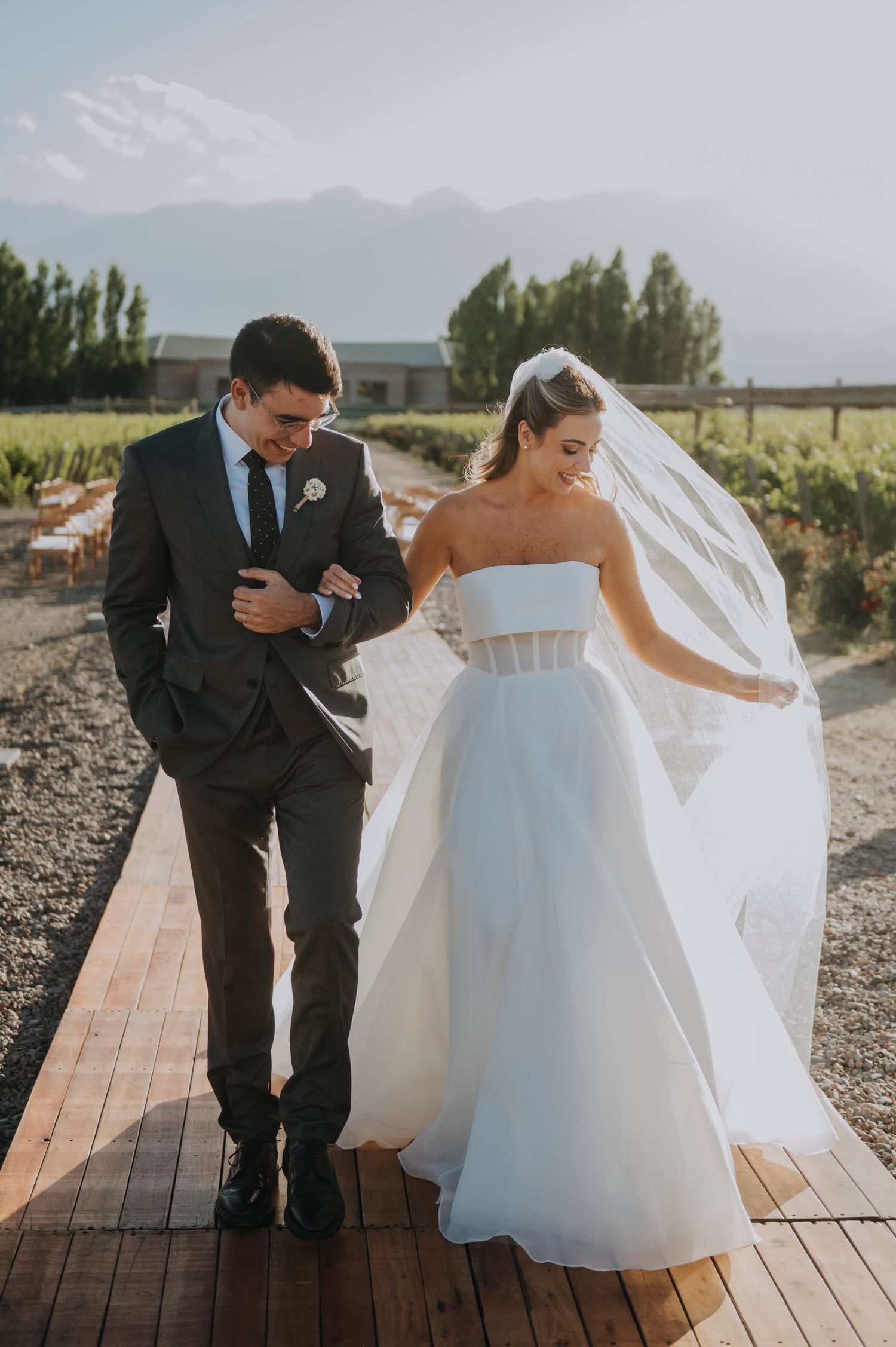 Bride in strapless white ballgown and groom in dark suit walk together on wooden path at vineyard wedding.