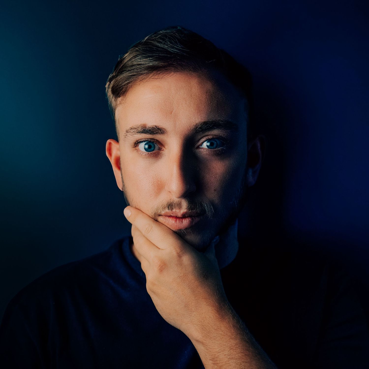 Young man with blue eyes resting chin on hand against dark blue background in dramatic portrait photography.
