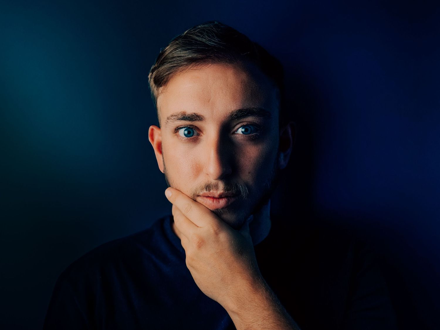 LEO FURIO Young man with light mustache resting chin on hand against dark blue background in moody portrair