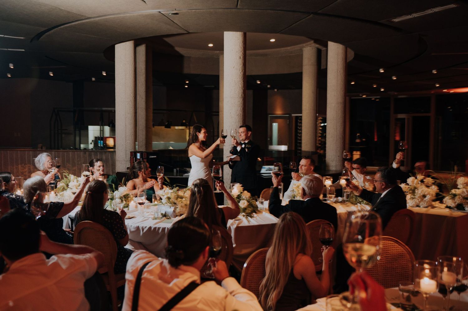 Wedding reception toast with guests raising glasses in a warmly lit elegant venue.