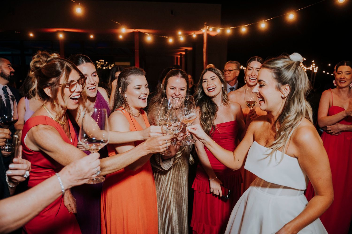 Bride celebrates with bridesmaids in red and orange dresses, toasting under string lights at reception.