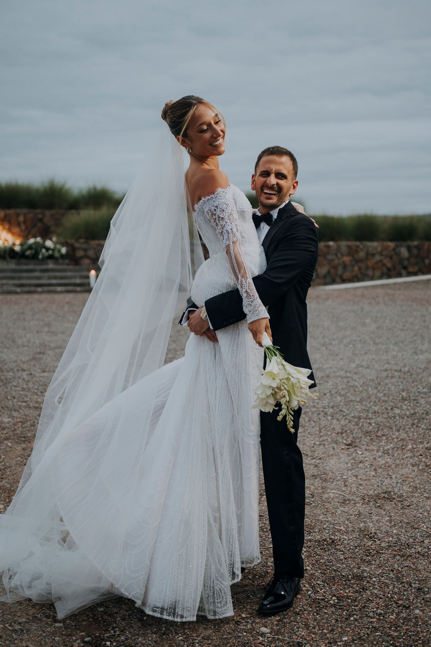 Smiling bride in off-shoulder lace wedding gown with veil poses with groom in black tuxedo.