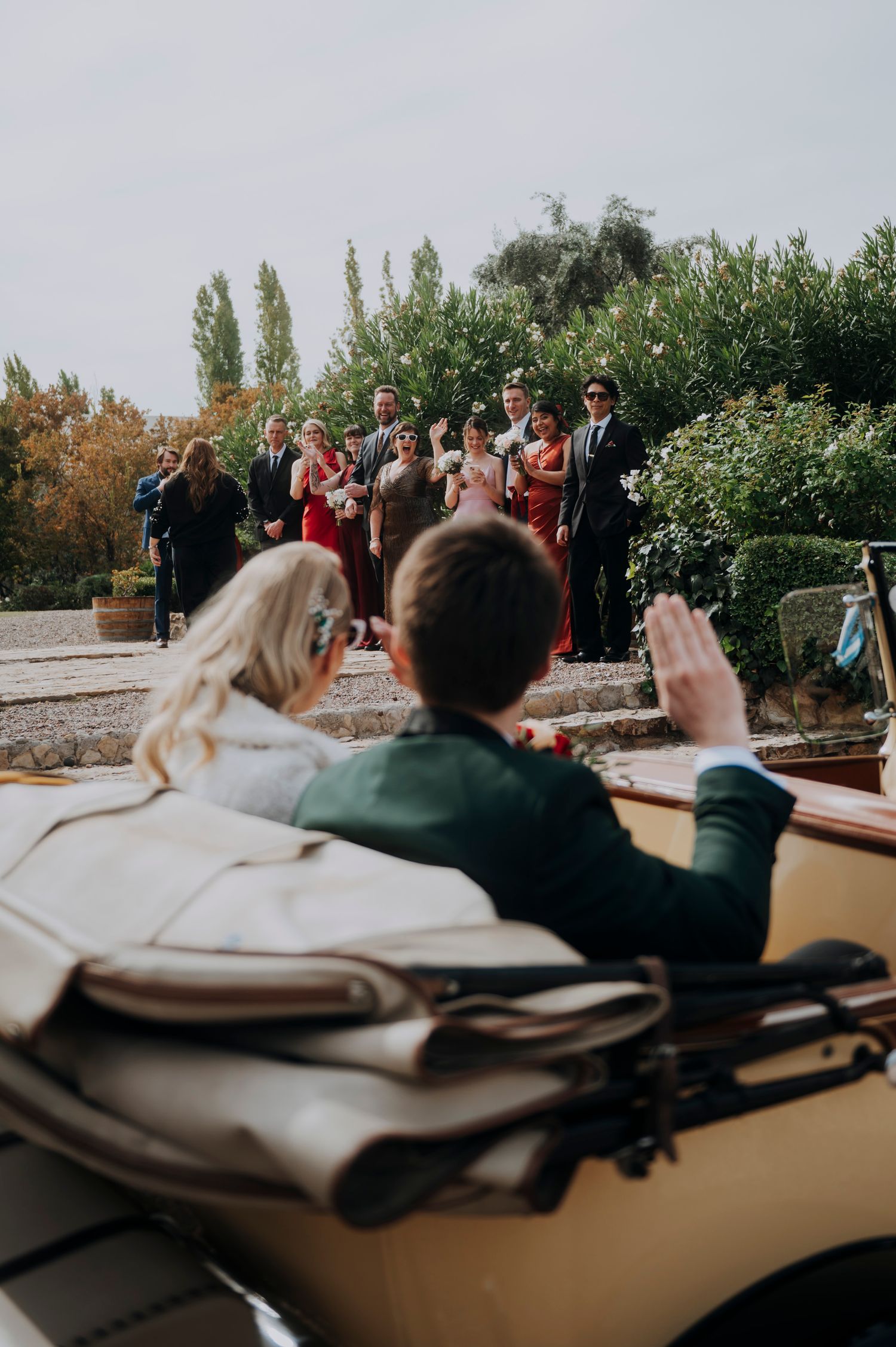 Newlyweds waving goodbye to wedding guests from vintage convertible car after ceremony.
