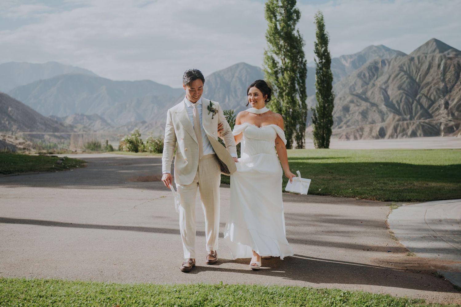 Bride and groom walking together on golf course path with scenic mountain backdrop.