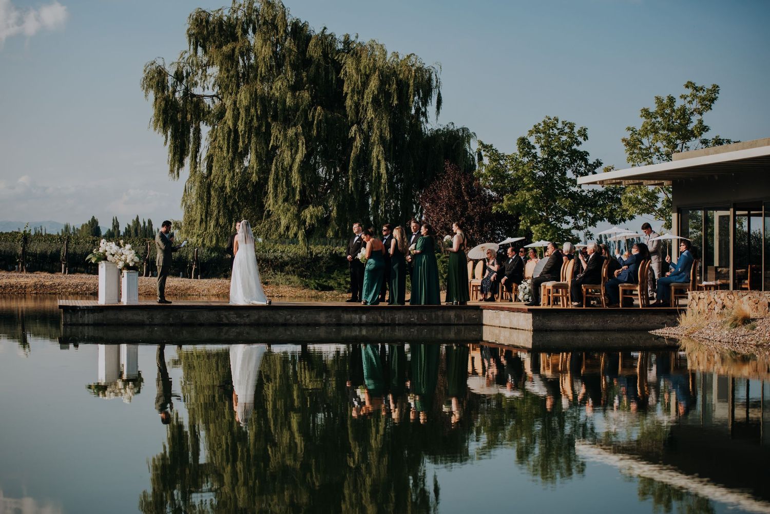 Outdoor wedding ceremony beside a reflective pond with guests gathered on a dock, willow tree backdrop.