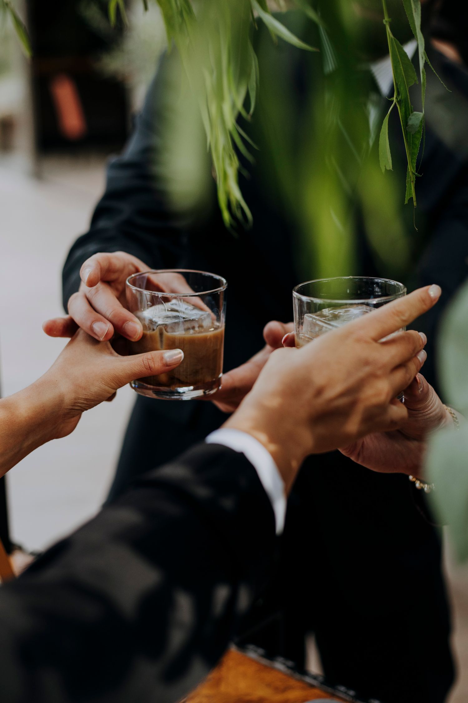 Wedding guests clink glasses of whiskey in a toast surrounded by lush greenery at reception.