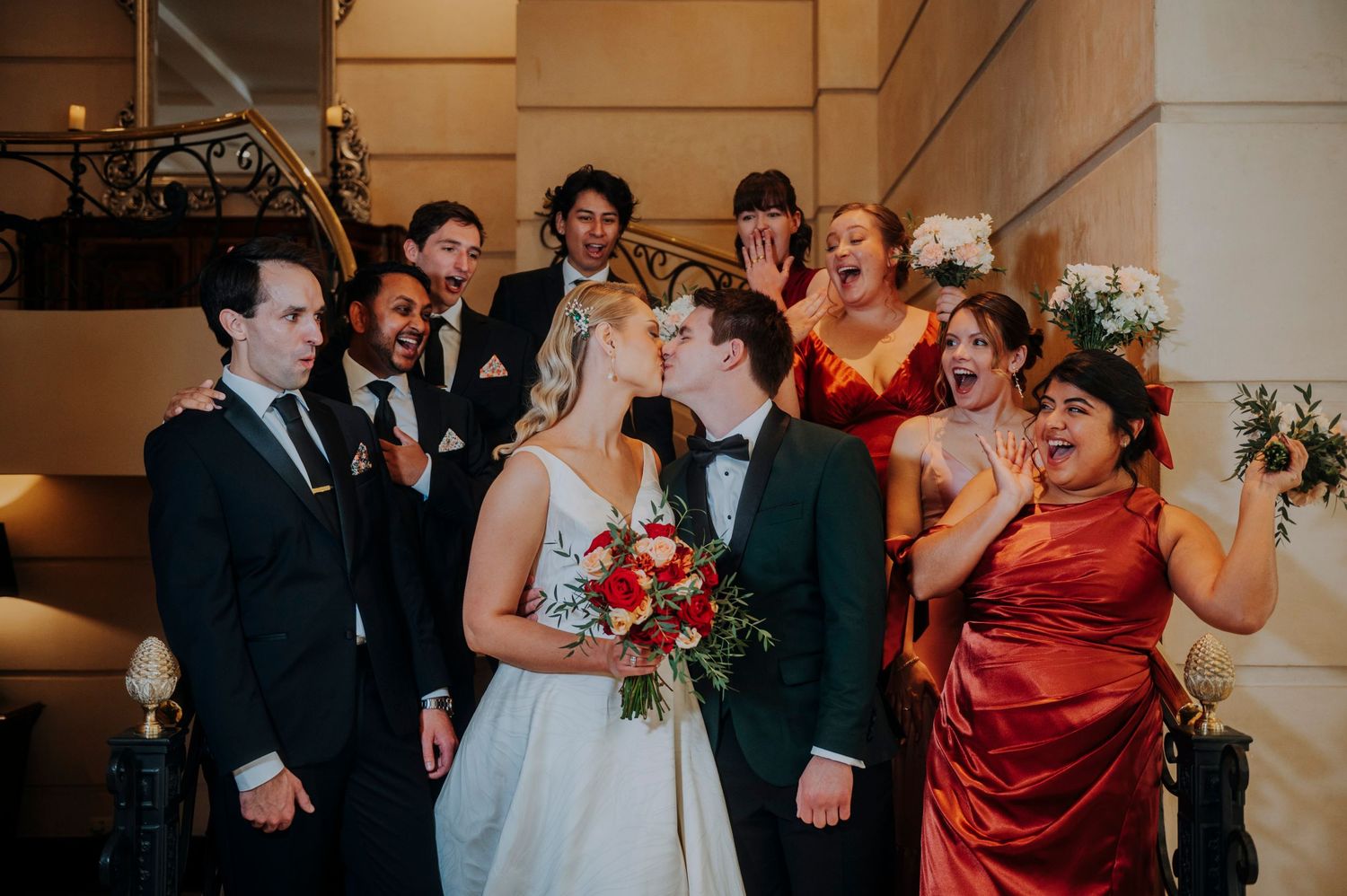 Bride and groom share a kiss surrounded by cheering wedding party on elegant staircase.