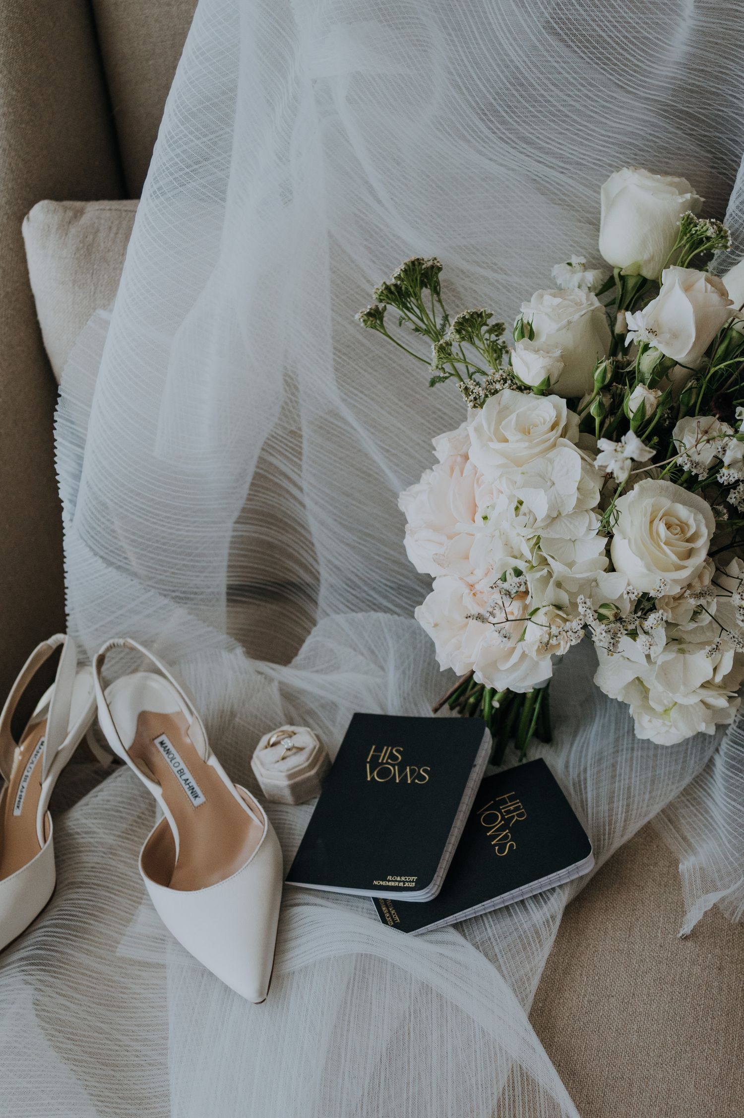 Elegant bridal flat lay with white heels, vow books, ring box, and white rose bouquet on a textured veil.