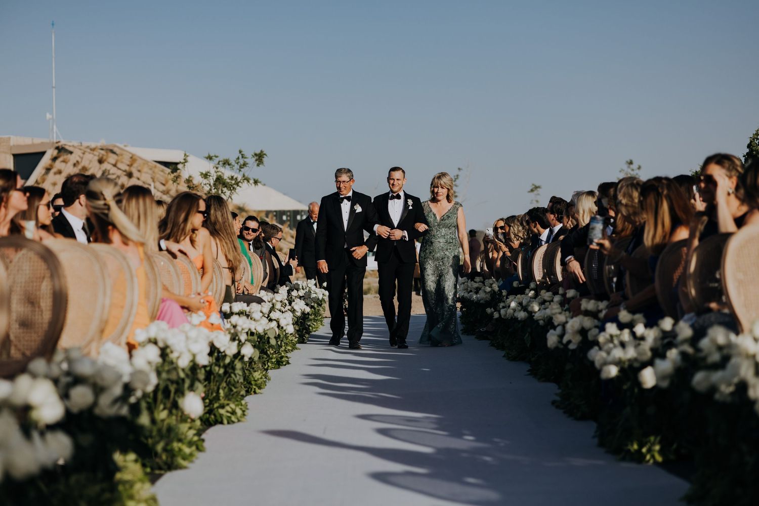Groom walks down the floral-lined aisle with two guests during an outdoor vineyard wedding ceremony.