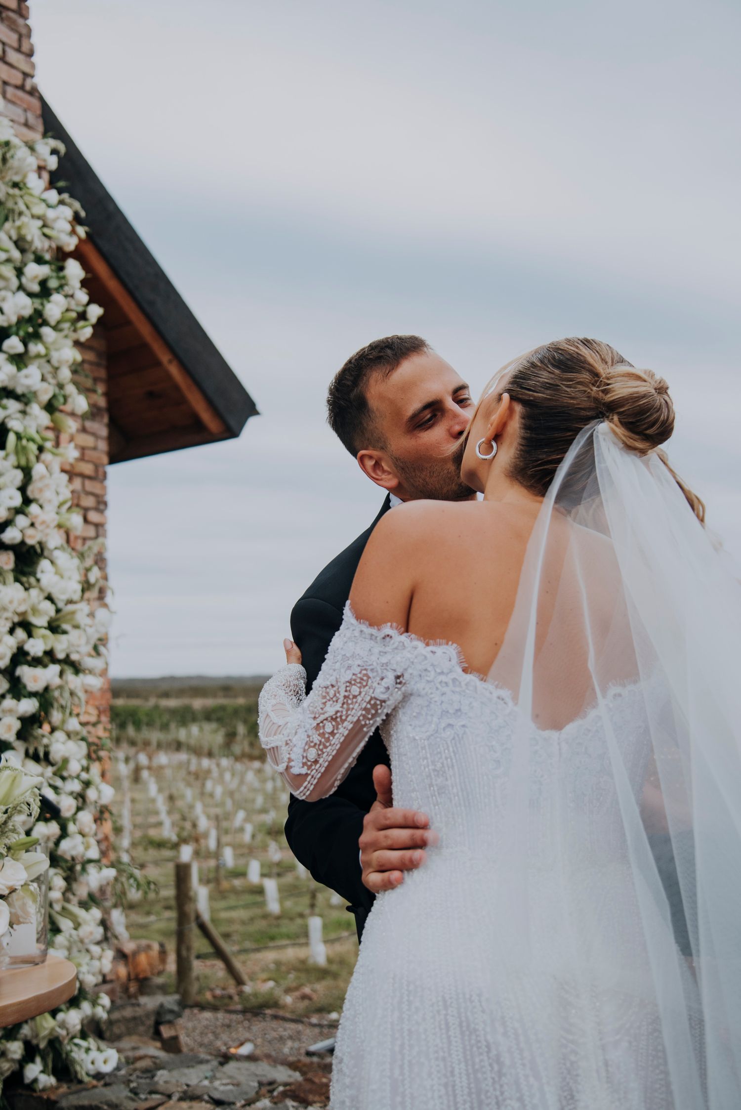 Groom kisses bride in lace off-shoulder wedding dress with veil at outdoor floral ceremony.