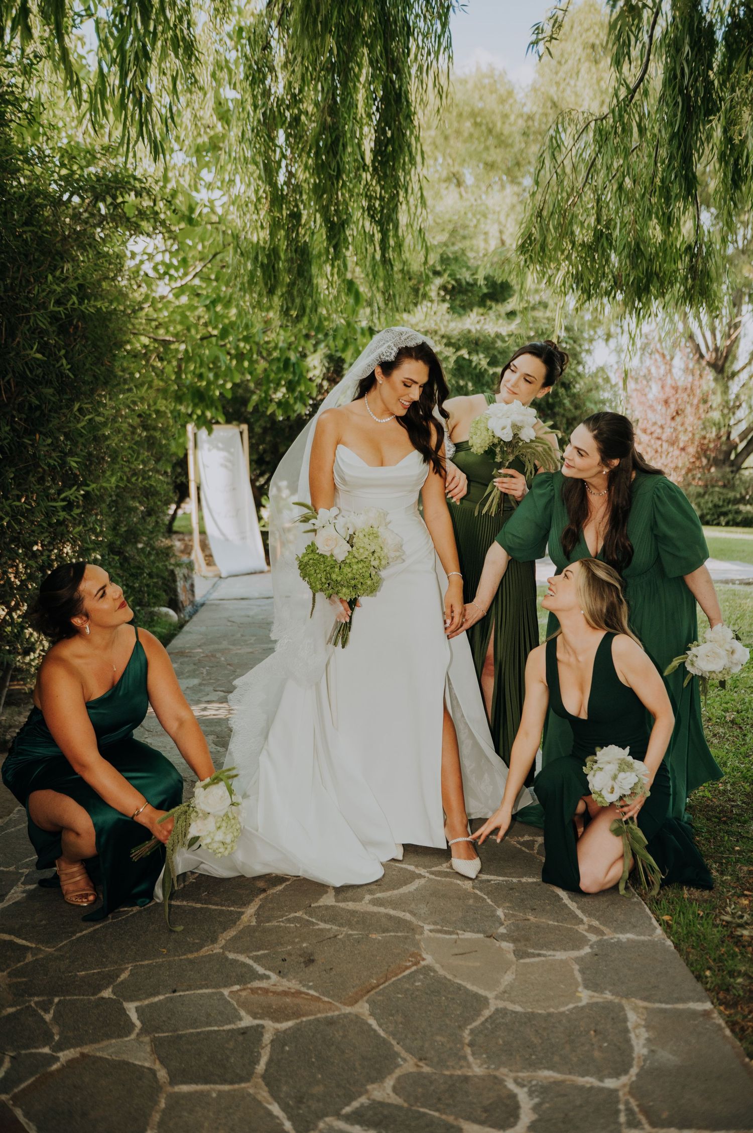 Bride in white gown with bridesmaids in emerald green dresses posing on stone path under willow tree.