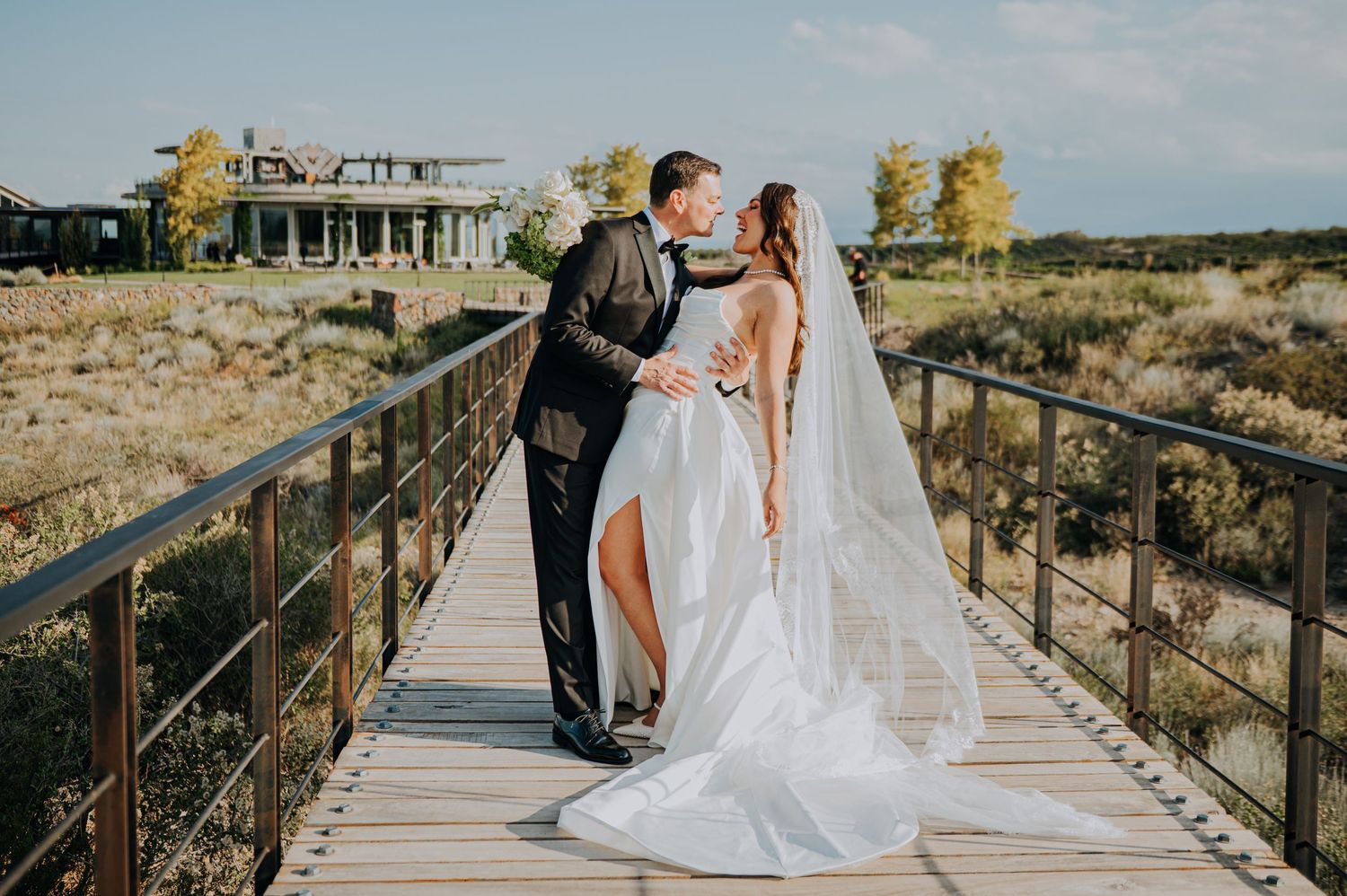 A bride and groom share a romantic kiss on a wooden bridge surrounded by scenic countryside on their wedding day.