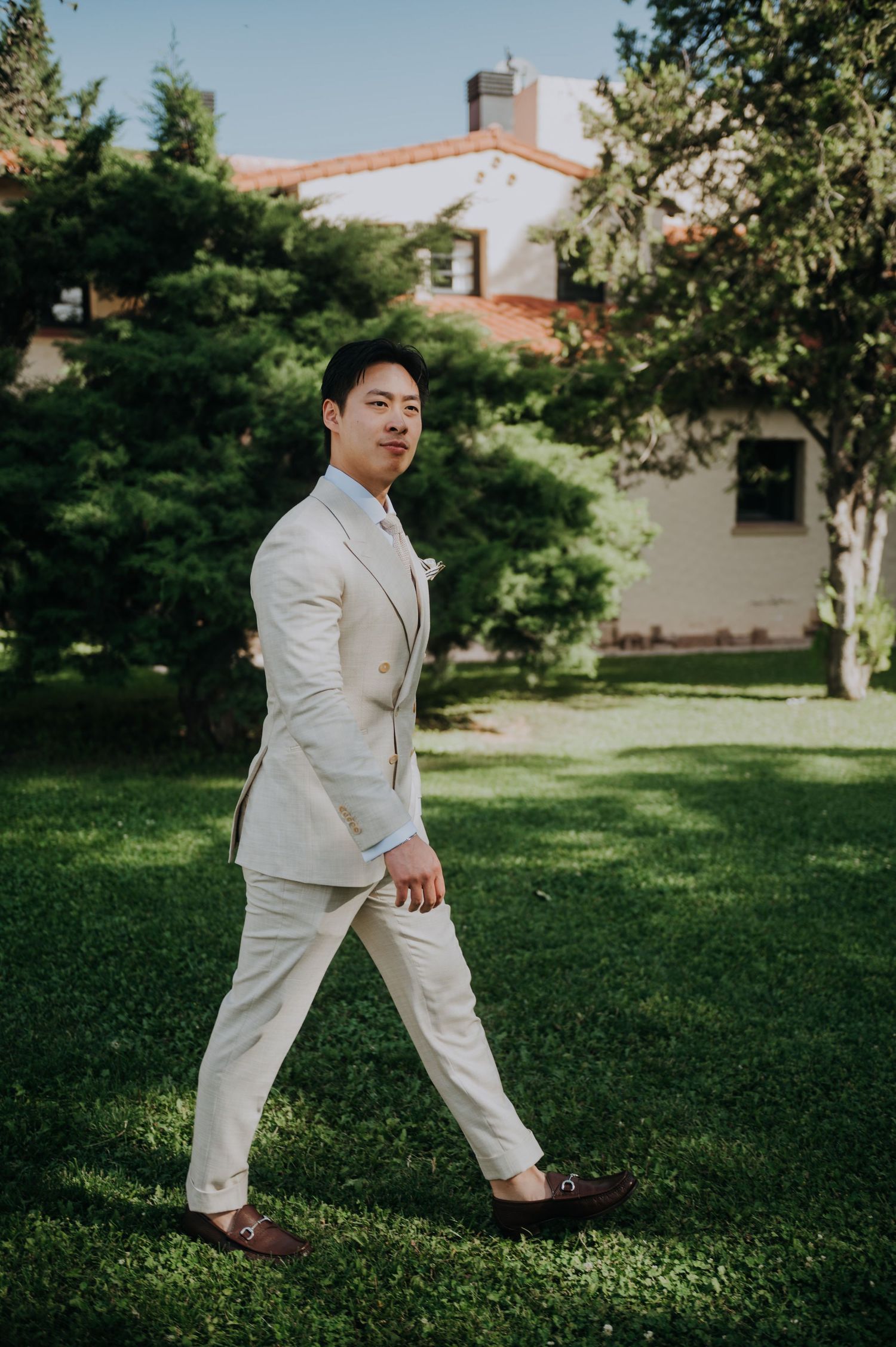 A well-dressed man in a cream suit walks across a lush green lawn with trees and a house in the background.