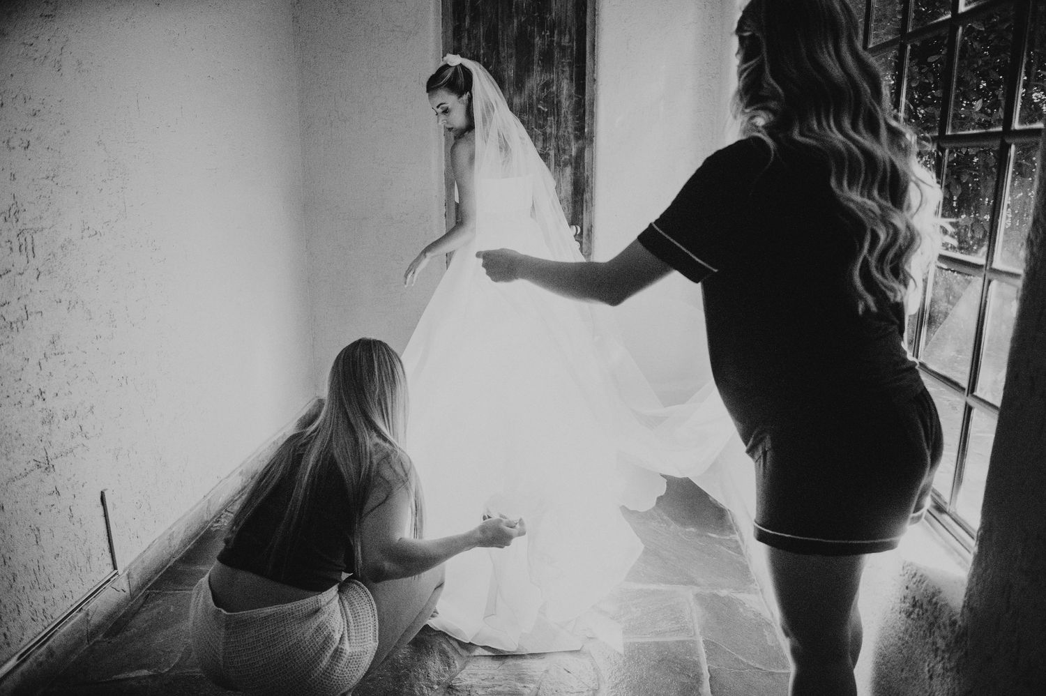 Black and white photo of bride in wedding dress and veil being assisted by two bridesmaids in a sunlit room.