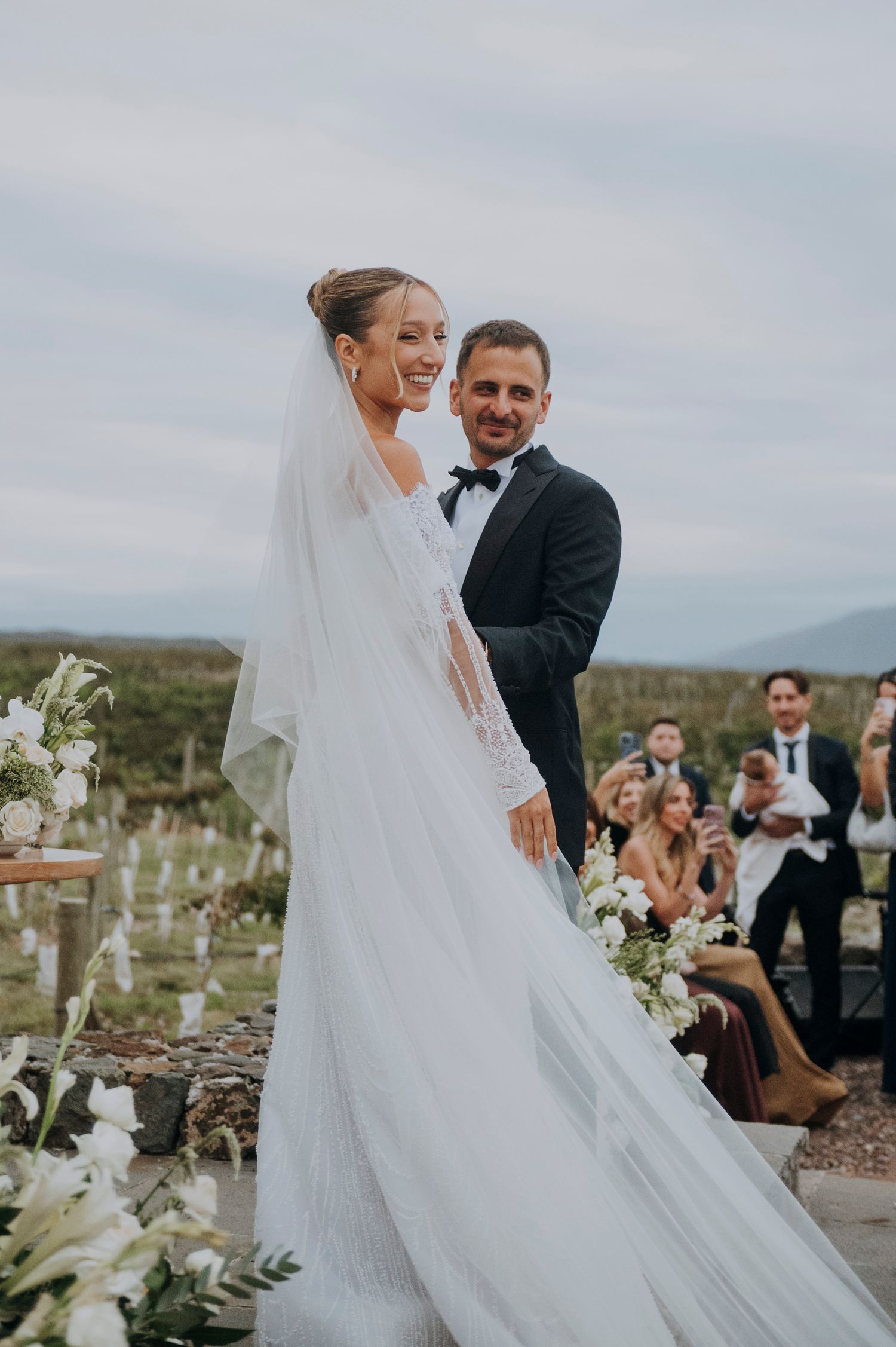 Bride and groom smile together at outdoor wedding ceremony with vineyard backdrop.