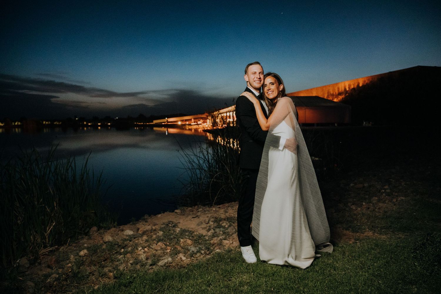 Newlyweds embrace by a serene lakeside at dusk, with glowing lights reflecting on the water behind them.