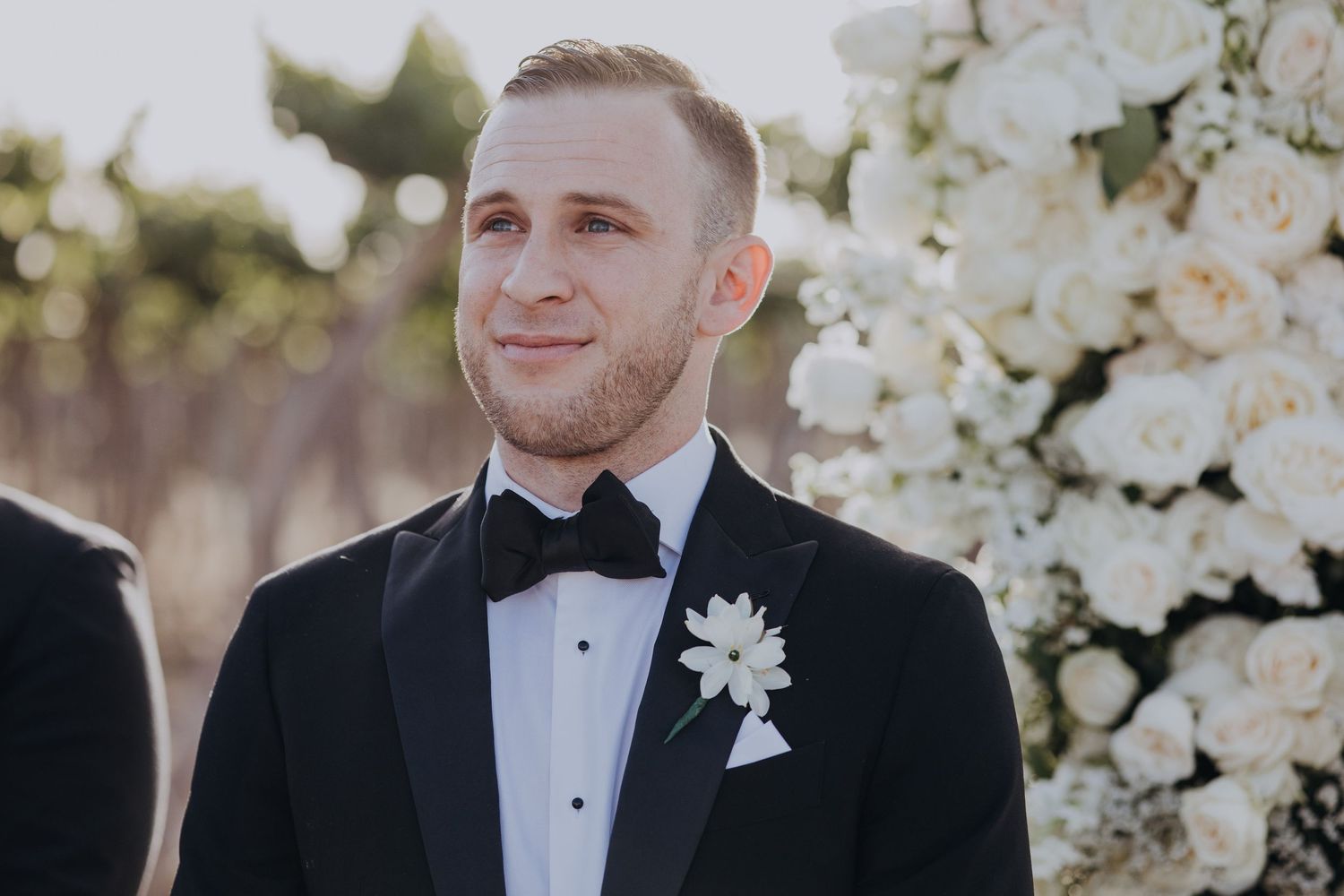 Groom in black tuxedo with white boutonniere smiling at outdoor wedding ceremony altar.