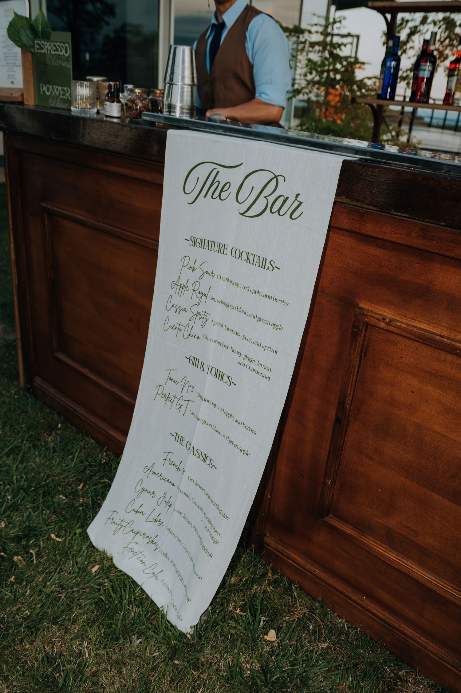 Elegant handwritten bar menu banner displayed on a rustic wooden bar at an outdoor wedding.