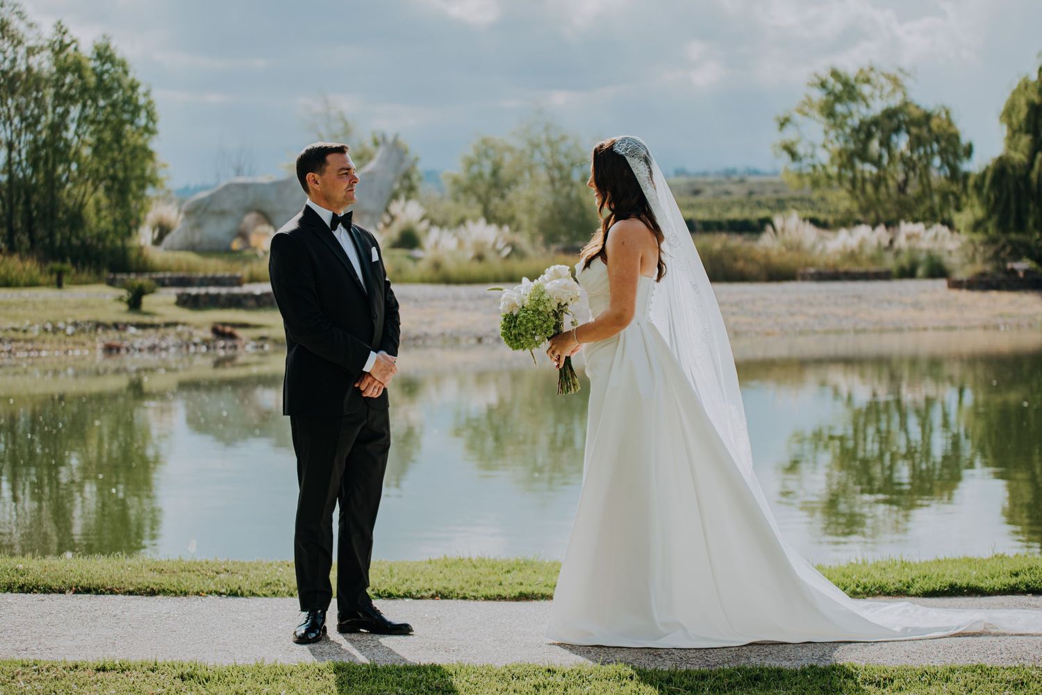 Bride and groom share a first look moment by a serene pond, bride in white gown with veil holding bouquet.