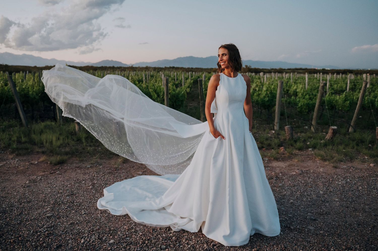 Bride in elegant white gown with flowing veil in vineyard at dusk.