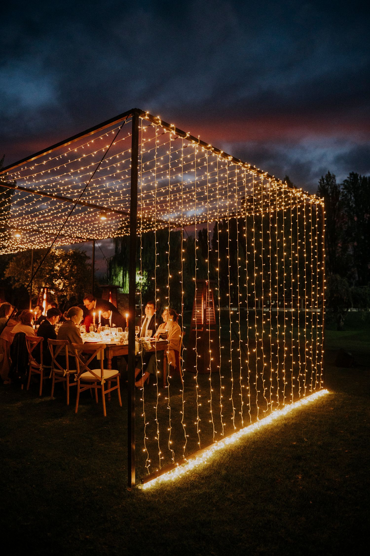 Outdoor wedding reception glows under curtain of fairy lights at twilight with dramatic sunset sky.