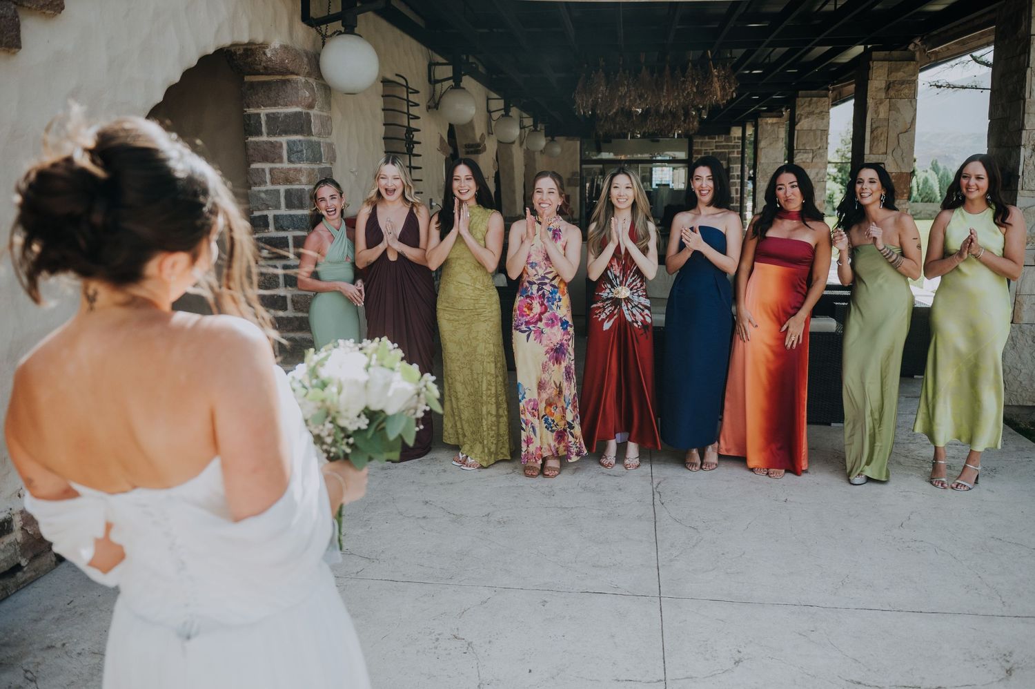 Bride reveals wedding dress to bridesmaids in colorful mismatched gowns, who react with joy and excitement.