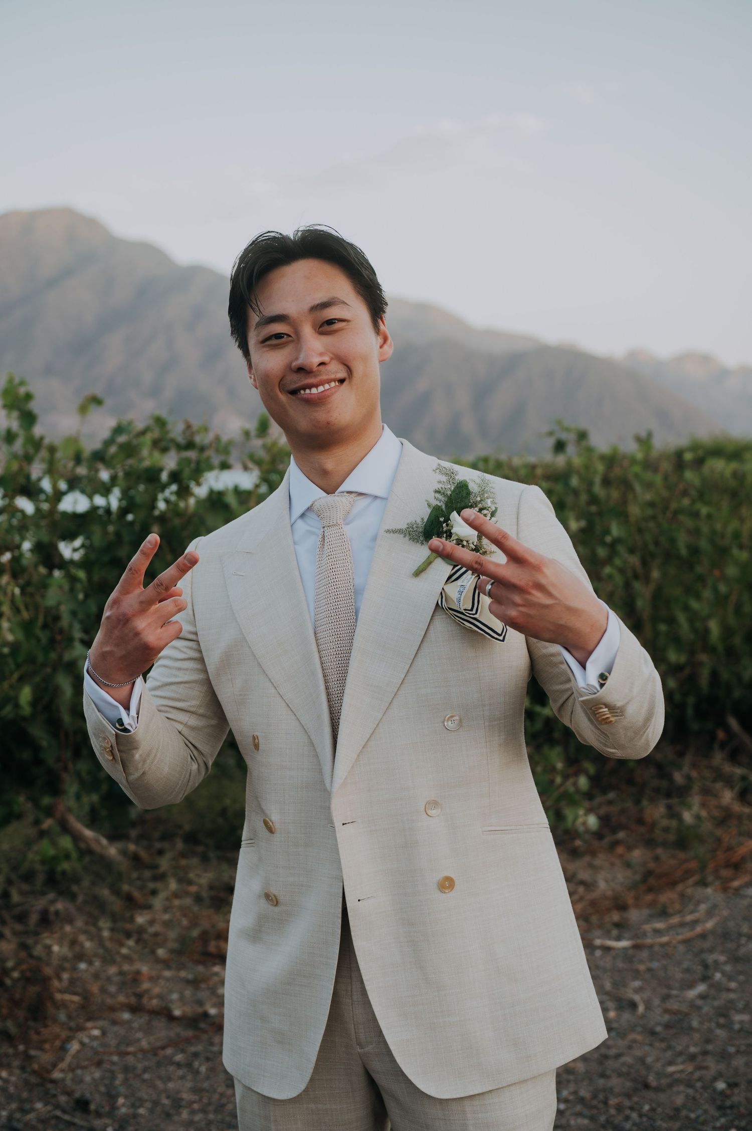 Smiling groom in cream suit making peace signs with mountain scenery behind him.